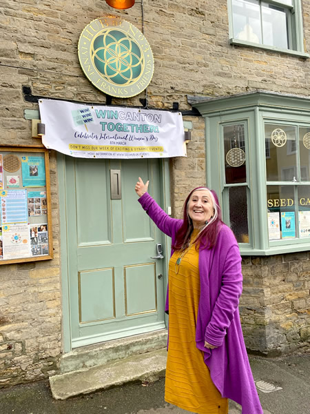 Lynne Franks, standing outside her SEED Hub, pointing at the Internation Women's Day sign over her door